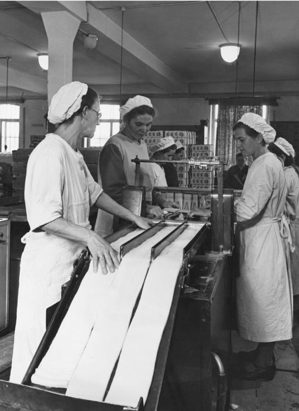 The image is a historical black and white photograph from the HARTMANN GROUP’s dermaplast active factory, taken between 1818 and 1918. It depicts three workers engaged in the production of medical supplies. They are dressed in period-appropriate work attire, complete with aprons and head coverings, and are handling long sheets of material on a production line.